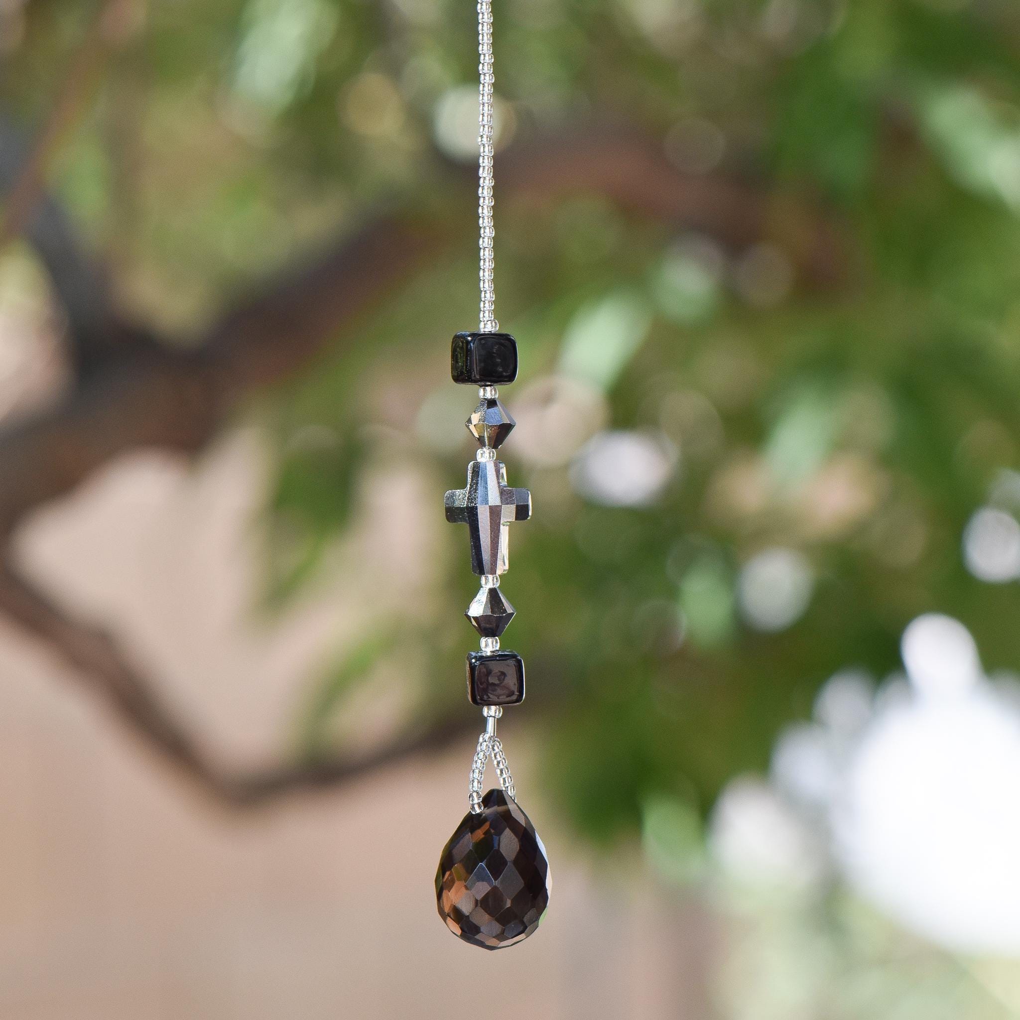 Gray Silver glass crystal cross beaded with two gray silver bicone beads and 2 Murano cubic black glass beads, hanging vertically against blurred background.