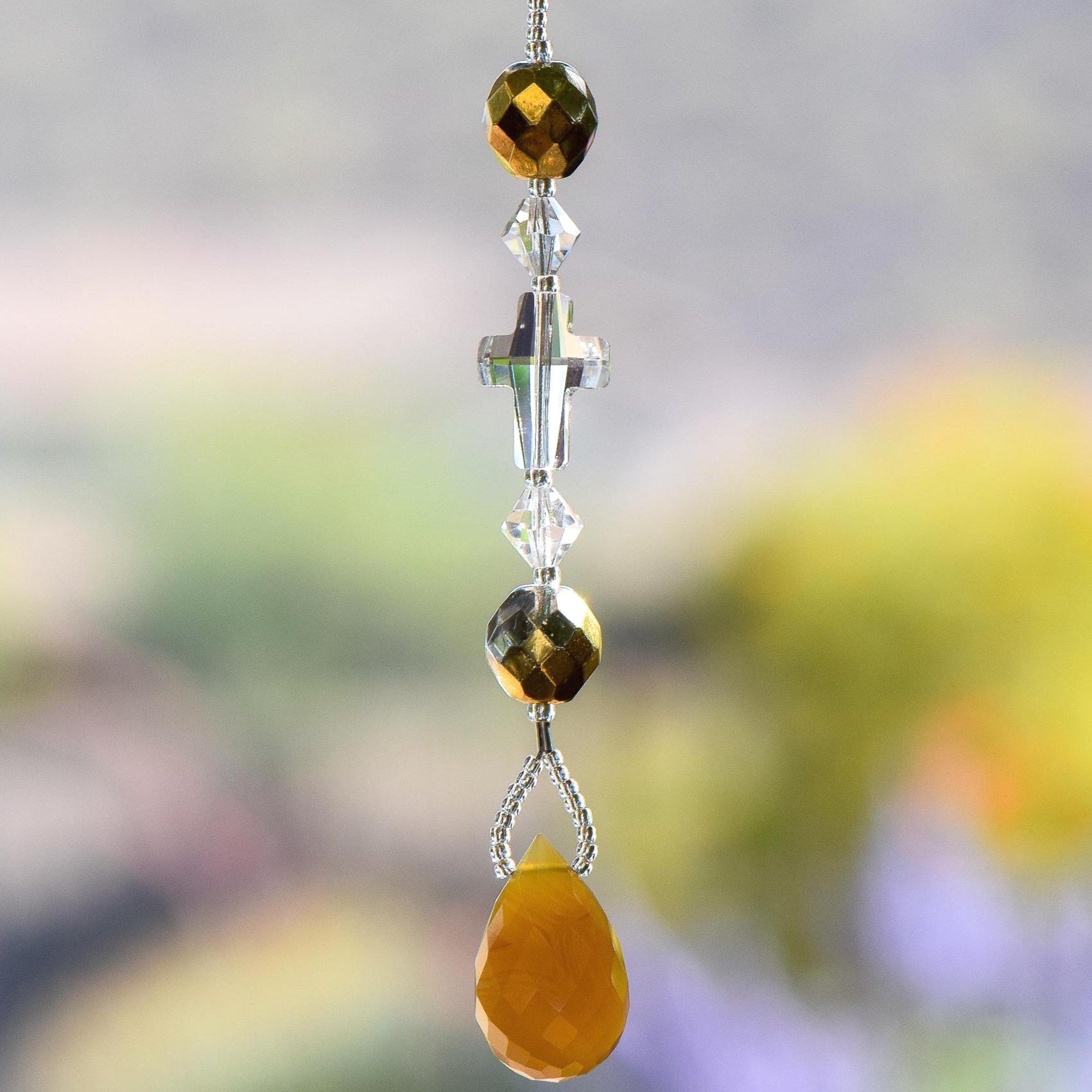 Glass crystal cross is beaded with other crystal beads and anchored by Carnelian Agate stone pendant, all hanging vertically against blurred background. For use as a small sun catcher in a window or hanging from the rear view mirror in a car.