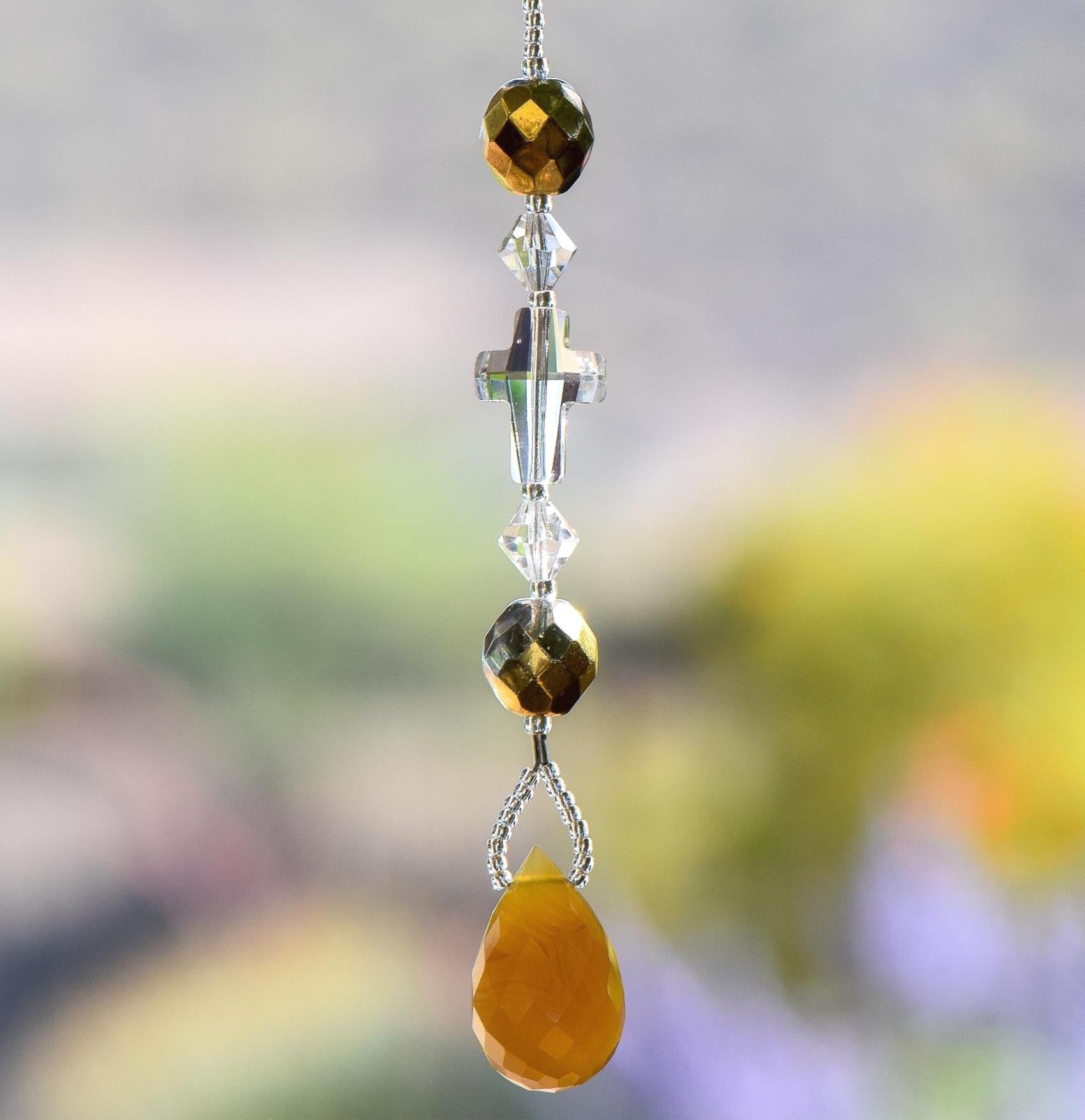 Glass crystal cross is beaded with other crystal beads and anchored by Carnelian Agate stone pendant, all hanging vertically against blurred background. For use as a small sun catcher in a window or hanging from the rear view mirror in a car.