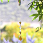 Glass crystal cross is beaded with other crystal beads and anchored by Carnelian Agate stone pendant, all hanging vertically from a hook in a tree, against blurred background. For use as a small sun catcher in a window or hanging from the rear view mirror in a car.