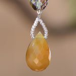 A faceted, teardrop-shaped carnelian agate stone pendant in shades of yellow and orange. The pendant is suspended by a string of small, clear beads below a larger, iridescent crystal bead. The background is a soft, blurred brown.