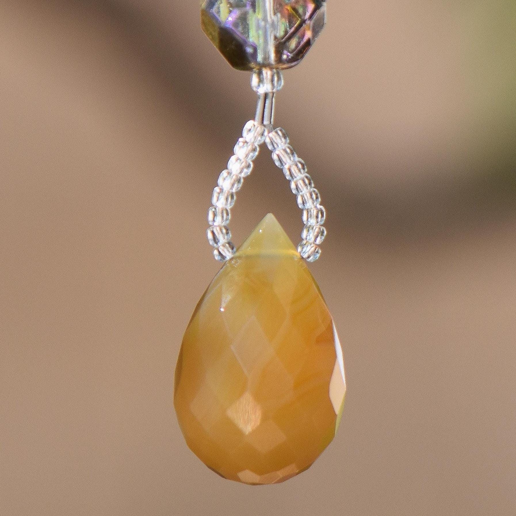 A faceted, teardrop-shaped carnelian agate stone pendant in shades of yellow and orange. The pendant is suspended by a string of small, clear beads below a larger, iridescent crystal bead. The background is a soft, blurred brown.