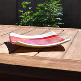 Square fused glass tray with vanilla cream colored base and a striped and streaky design of red, orange and white glass. Tray is 6&quot; square with upturned corners. Sitting on a wood table in the sunlight. Camera is viewing the tray from the side, at an angle. There is a plant in the background behind the wood table.