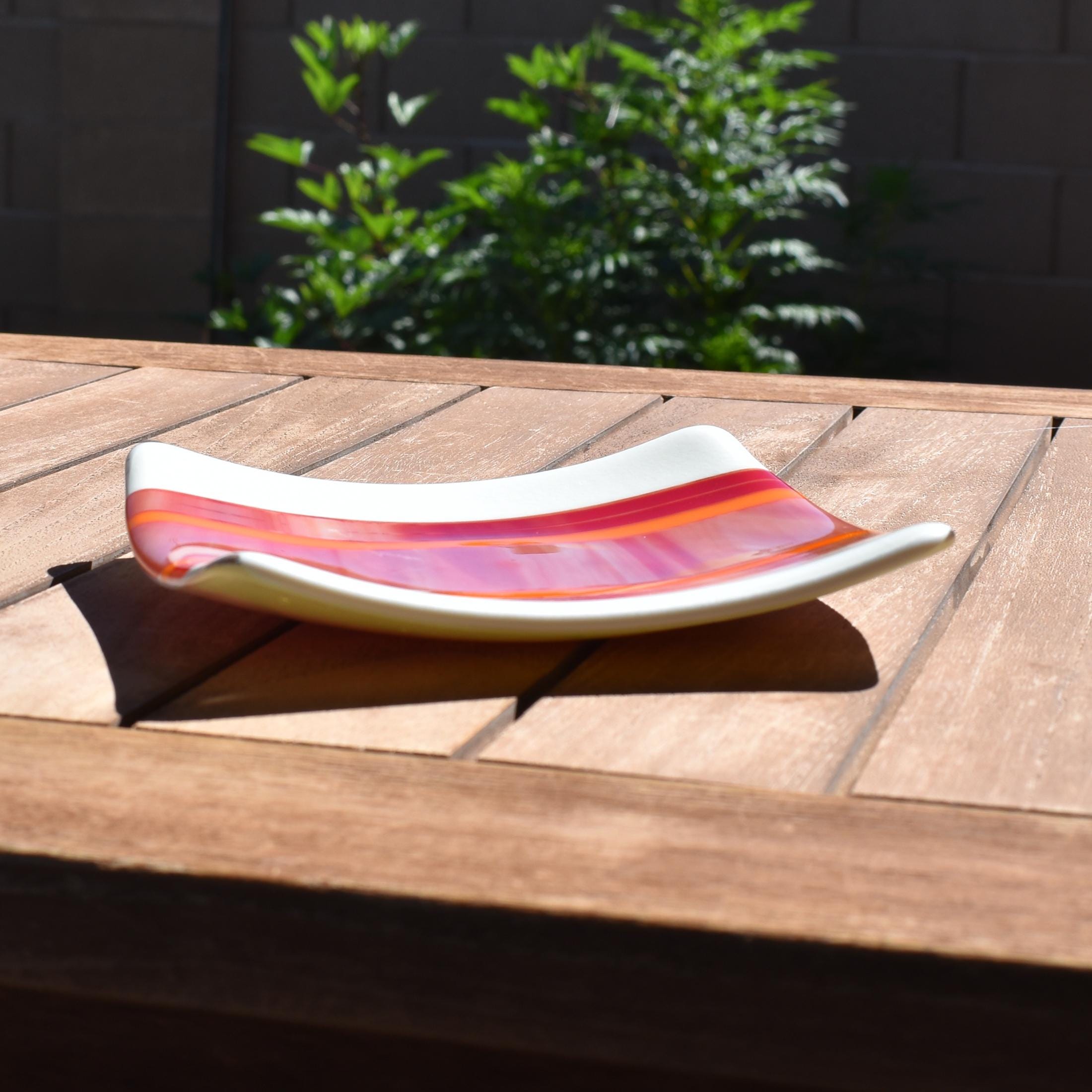 Square fused glass tray with vanilla cream colored base and a striped and streaky design of red, orange and white glass. Tray is 6&quot; square with upturned corners. Sitting on a wood table in the sunlight. Camera is viewing the tray from the side, at an angle. There is a plant in the background behind the wood table.