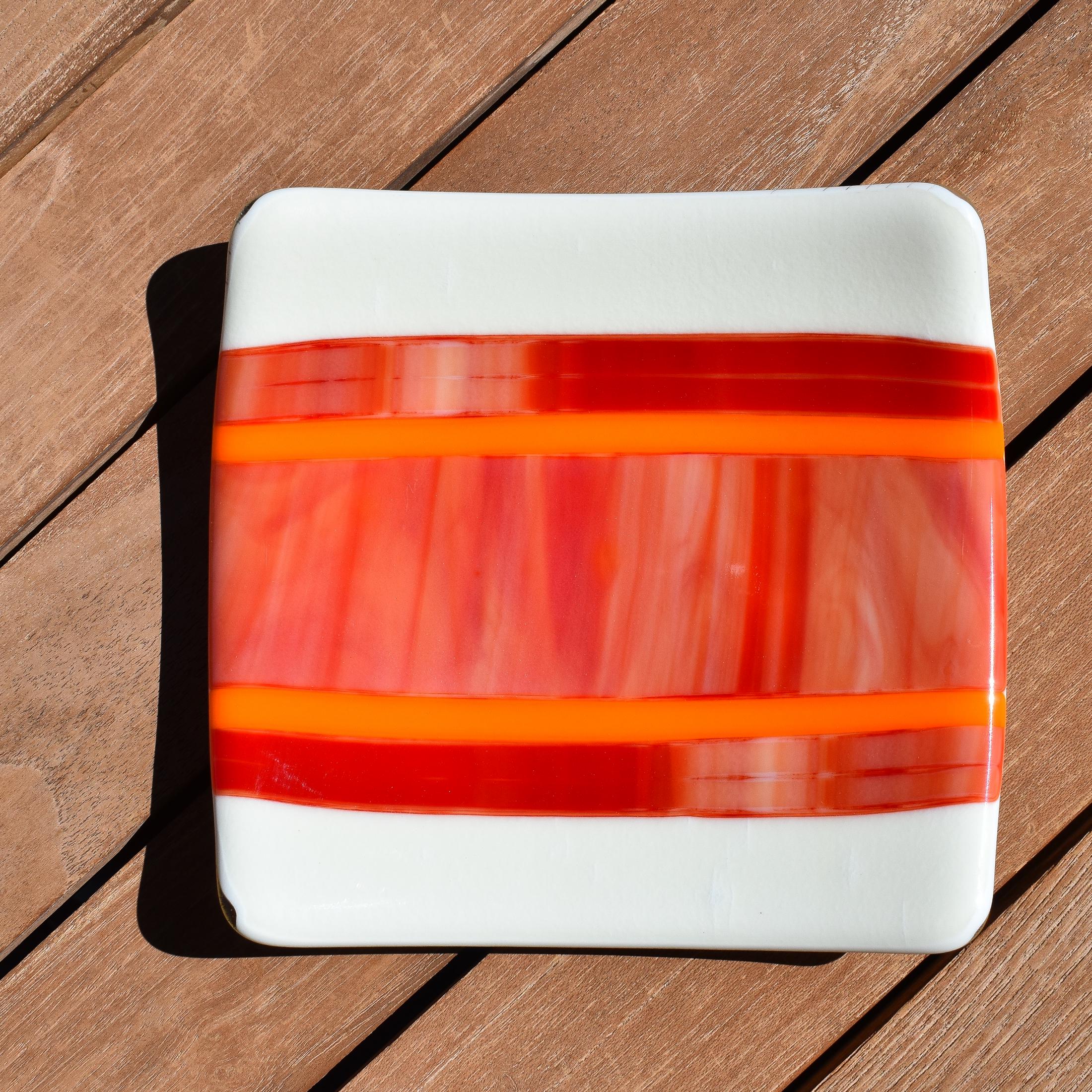 Square fused glass tray with vanilla cream colored base and a striped and streaky design of red, orange and white glass. Tray is 6&quot; square with upturned corners. Sitting on a wood table in the sunlight. View is from the top down.