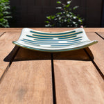 Square glass dish, viewed from the front, on a slatted wood table. The dish has a base color of vanilla cream with abstract design consisting of lines of peacock green.