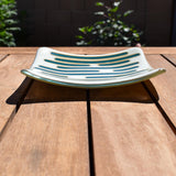 Square glass dish, viewed from the front, on a slatted wood table. The dish has a base color of vanilla cream with abstract design consisting of lines of peacock green.