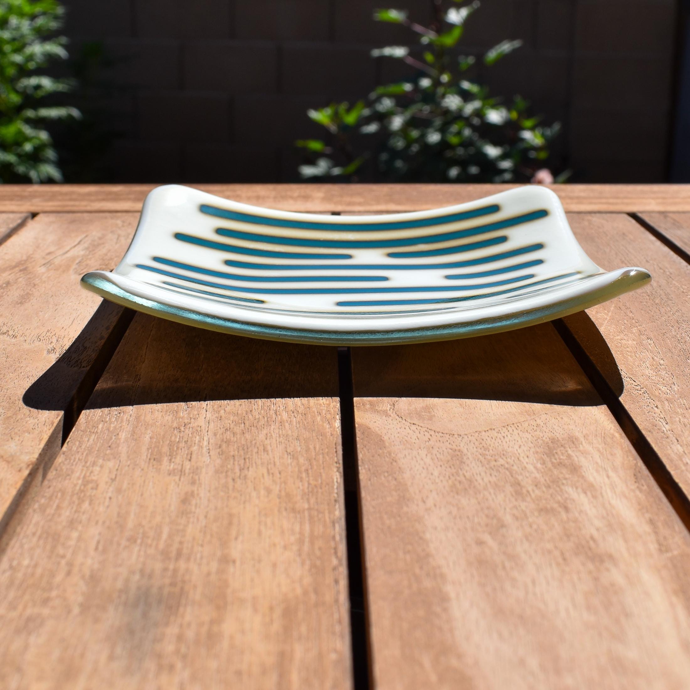 Square glass dish, viewed from the front, on a slatted wood table. The dish has a base color of vanilla cream with abstract design consisting of lines of peacock green.