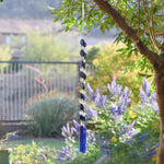 A blue glass wind chime hanging from a tree branch. The chime features two strings of dark blue lapis lazuli stone beads and clear glass beads, leading to two rectangular blue & plum purple glass pieces at the bottom. The two strings are joined at the top where the wire is hanging from a hook. The background shows a blurred view of green foliage and a house.