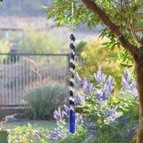 A blue glass wind chime hanging from a tree branch. The chime features two strings of dark blue lapis lazuli stone beads and clear glass beads, leading to two rectangular blue & plum purple glass pieces at the bottom. The two strings are joined at the top where the wire is hanging from a hook. The background shows a blurred view of green foliage and a house.