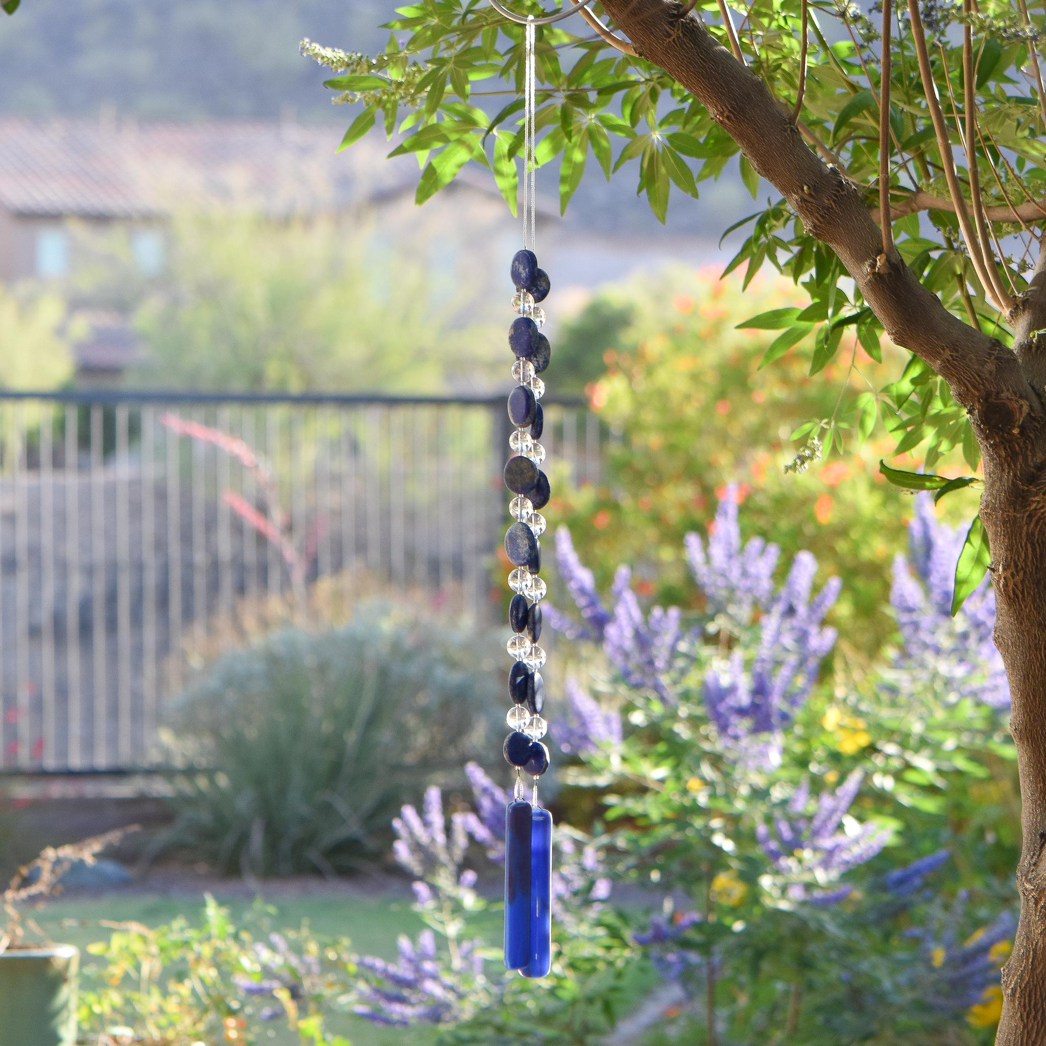 A blue glass wind chime hanging from a tree branch. The chime features two strings of dark blue lapis lazuli stone beads and clear glass beads, leading to two rectangular blue & plum purple glass pieces at the bottom. The two strings are joined at the top where the wire is hanging from a hook. The background shows a blurred view of green foliage and a house.