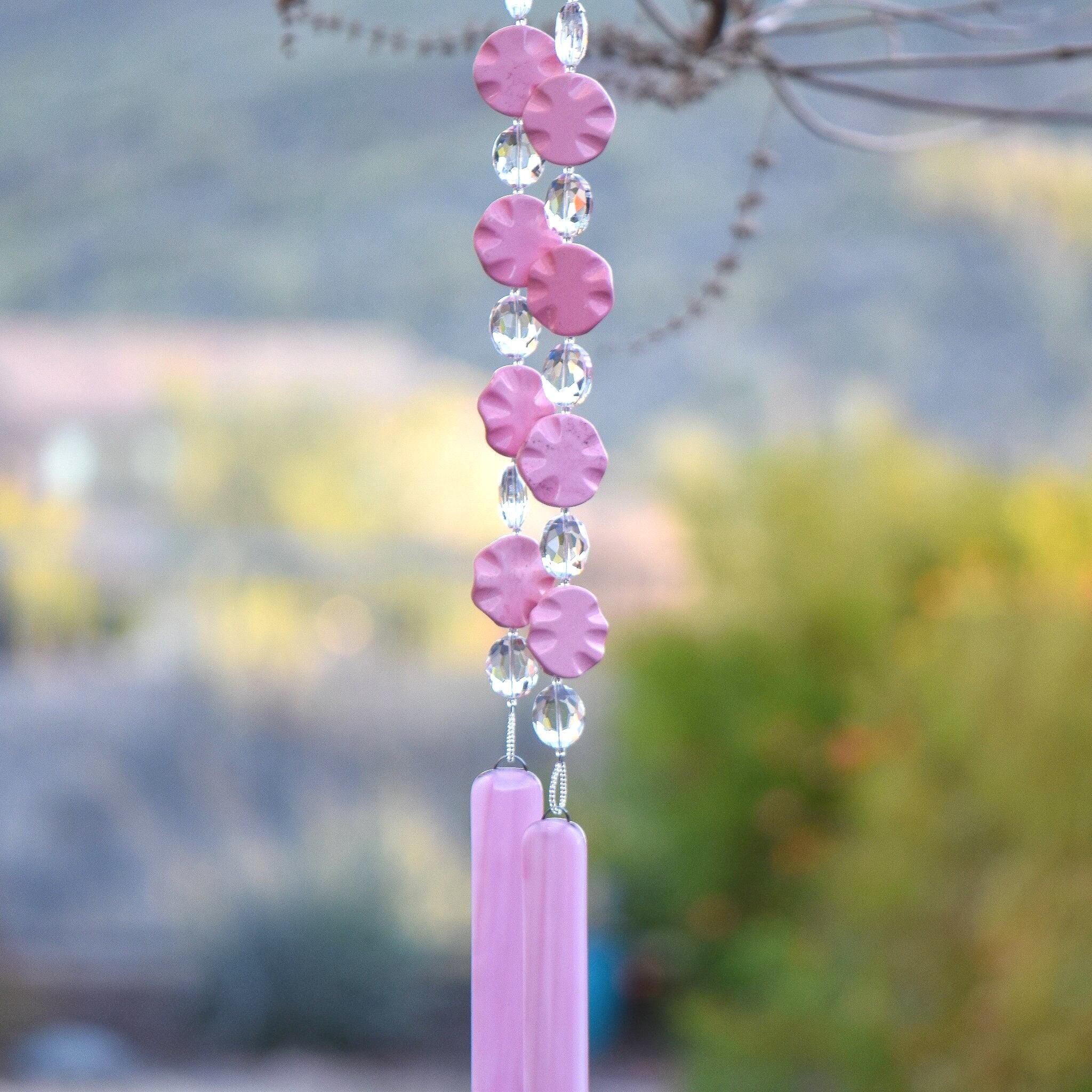 Pink magnesite stone beads shaped as wavy flat rounds are strung with glass crystal ovals and anchored by two pieces of pink fused glass. Hanging from tree. Blurred foliage background.