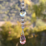 Small sun catcher on stainless steel wire hanging vertically in front of blurred landscape background. A large glass crystal bead is flanked by smaller beads with glass &quot;crackle&quot; and anchored by a very large faceted stone pendant.