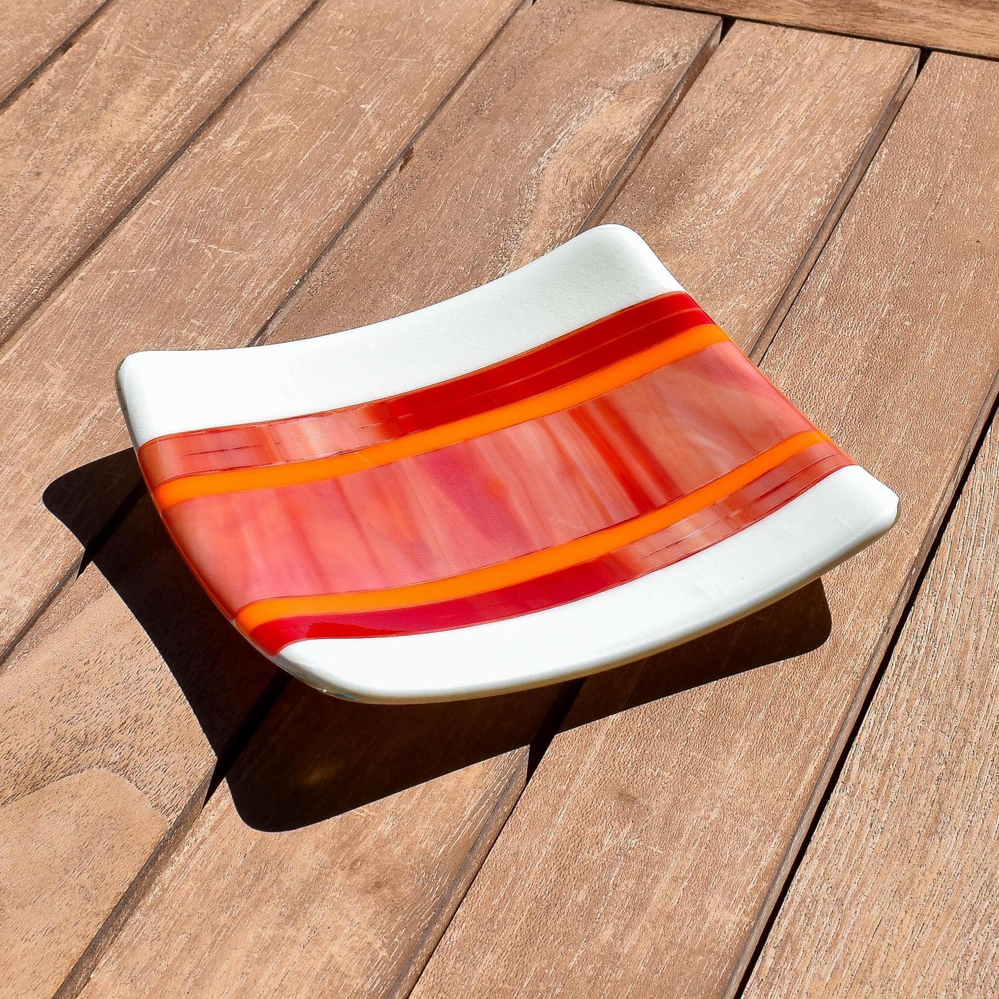 Square fused glass tray with vanilla cream colored base and a striped and streaky design of red, orange and white glass. Tray is 6&quot; square with upturned corners. Sitting on a wood table in the sunlight.