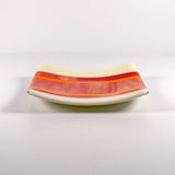 Square fused glass tray with vanilla cream colored base and a striped and streaky design of red, orange and white glass. Tray is 6&quot; square with upturned corners. Sitting on a white surface.