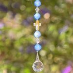 Beaded sun catcher hanging vertically against blurred green foliage background. Sun catcher has 4 light blue round glass beads, two bicone crystal beads. Center is a faceted glass cross bead with Crystal AB finish. Anchored by faceted glass round prism.