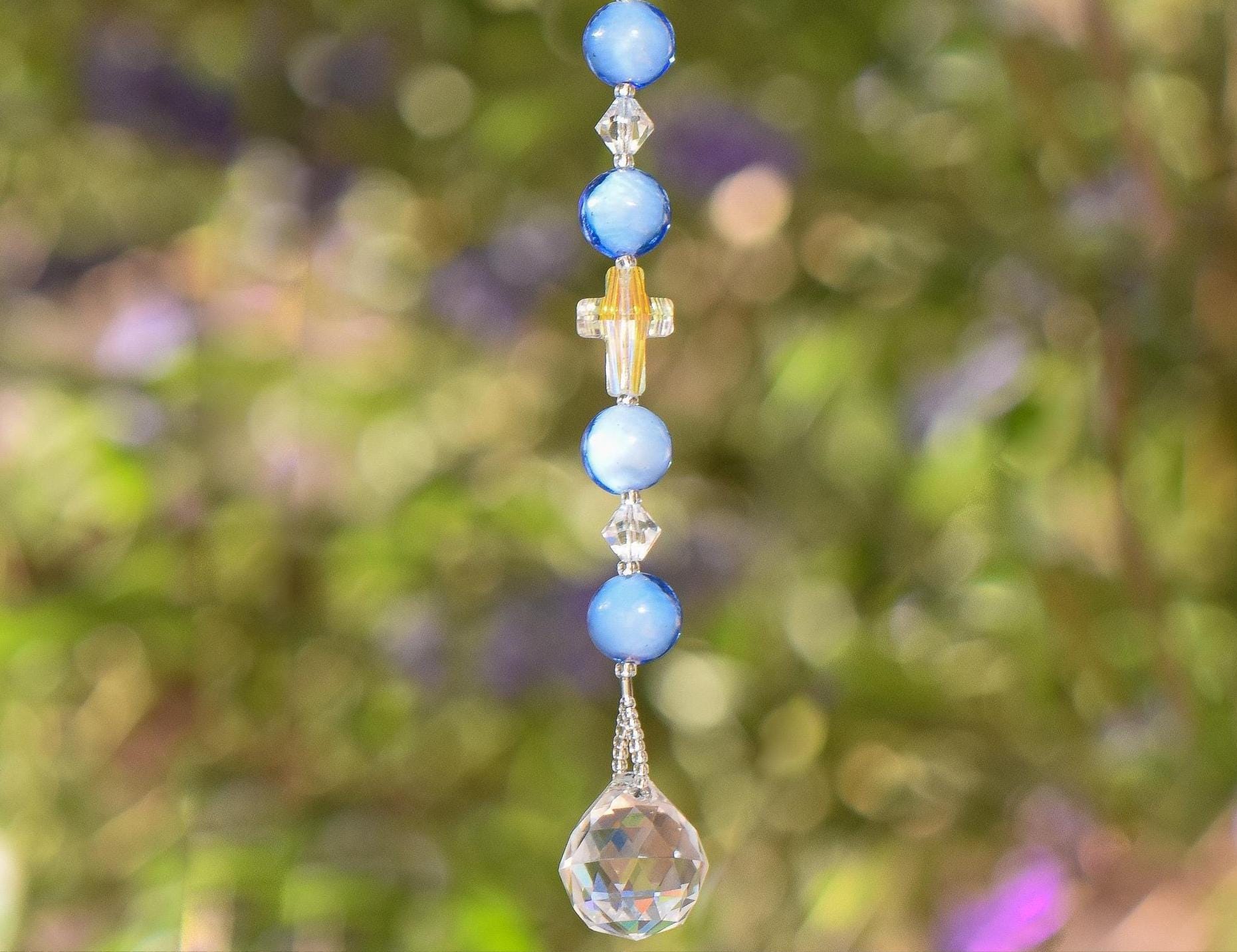Beaded sun catcher hanging vertically against blurred green foliage background. Sun catcher has 4 light blue round glass beads, two bicone crystal beads. Center is a faceted glass cross bead with Crystal AB finish. Anchored by faceted glass round prism.