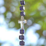 Faceted glass crystal cross bead strung on stainless steel wire, flanked by six purple murano glass cubic beads, all hanging vertically against blurred foliage background.