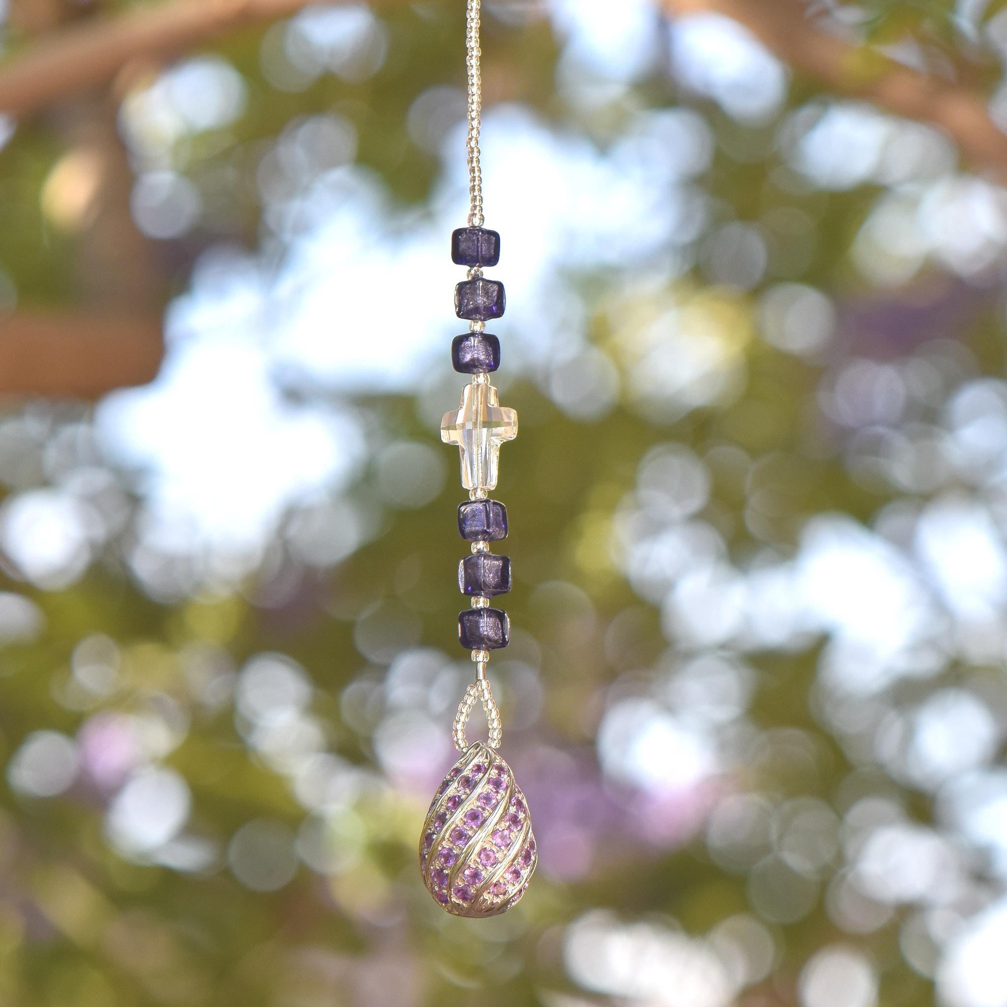 Small 9&quot; beaded sun catcher hanging vertically against blurred foliage background. Sun catcher is made with six Murano glass beads in purple with silver foil flanking a faceted glass crystal cross, anchored by a very large sterling silver pendant made with swirls of purple crystals.