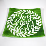 A square green glass plate with a white wreath design. The wreath has white leaves and the words &#39;Joy to the World&#39; in white script inside the wreath.