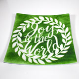 A square green glass plate with a white wreath design. The wreath has white leaves and the words &#39;Joy to the World&#39; in white script inside the wreath.