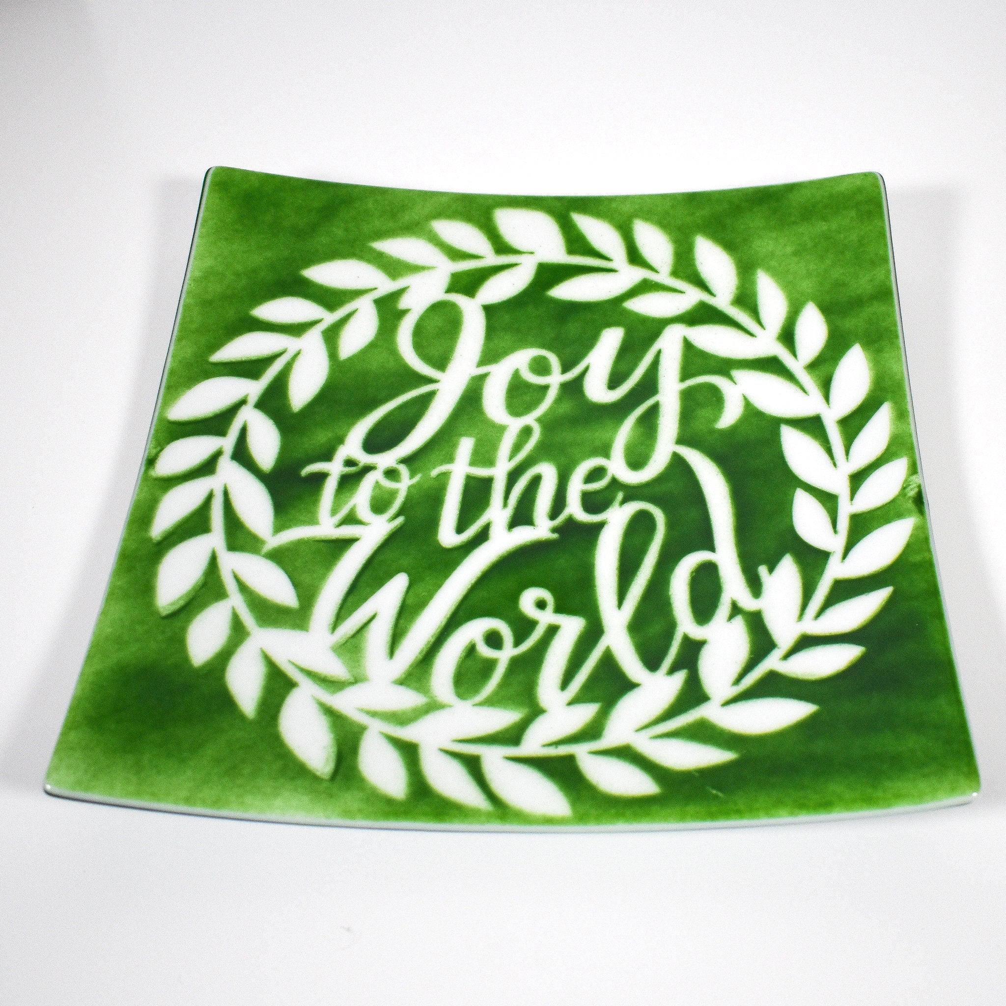 A square green glass plate with a white wreath design. The wreath has white leaves and the words &#39;Joy to the World&#39; in white script inside the wreath.