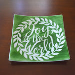 A square green glass dish with a white wreath design and the words &#39;Joy to the World&#39; in white lettering. On wood background.