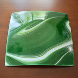 Backside of a square green glass plate showing white swirl design on sparkly aventurine green glass. The plate has a glossy finish and a subtle sparkle.