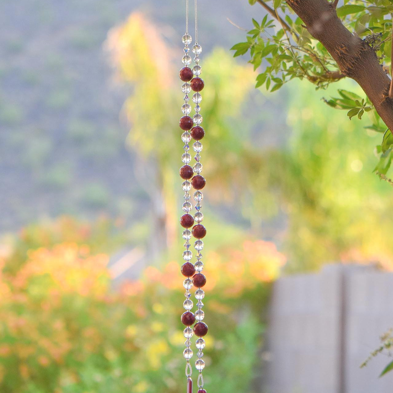 Large cranberry red jade stones beaded with glass and crystals, anchored by two pieces of dark cranberry pink fused glass, hanging vertically from a tree.