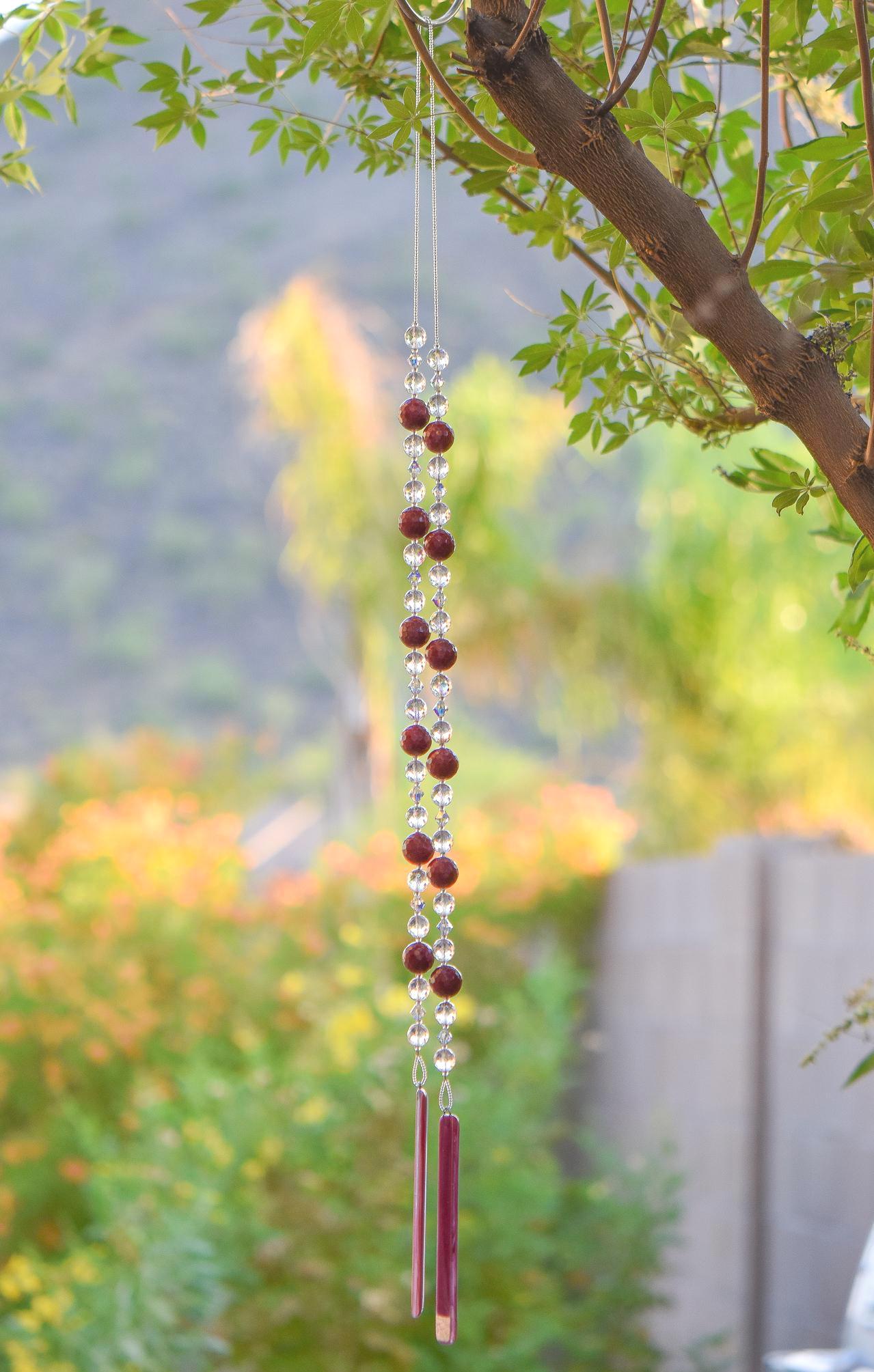 Large cranberry red jade stones beaded with glass and crystals, anchored by two pieces of dark cranberry pink fused glass, hanging vertically from a tree.