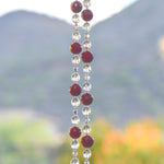 Two strands of beaded wiring hanging vertically against landscape background. Each wire has dark pinkish red jade stone beads flanked by clear glass and glass crystal.
