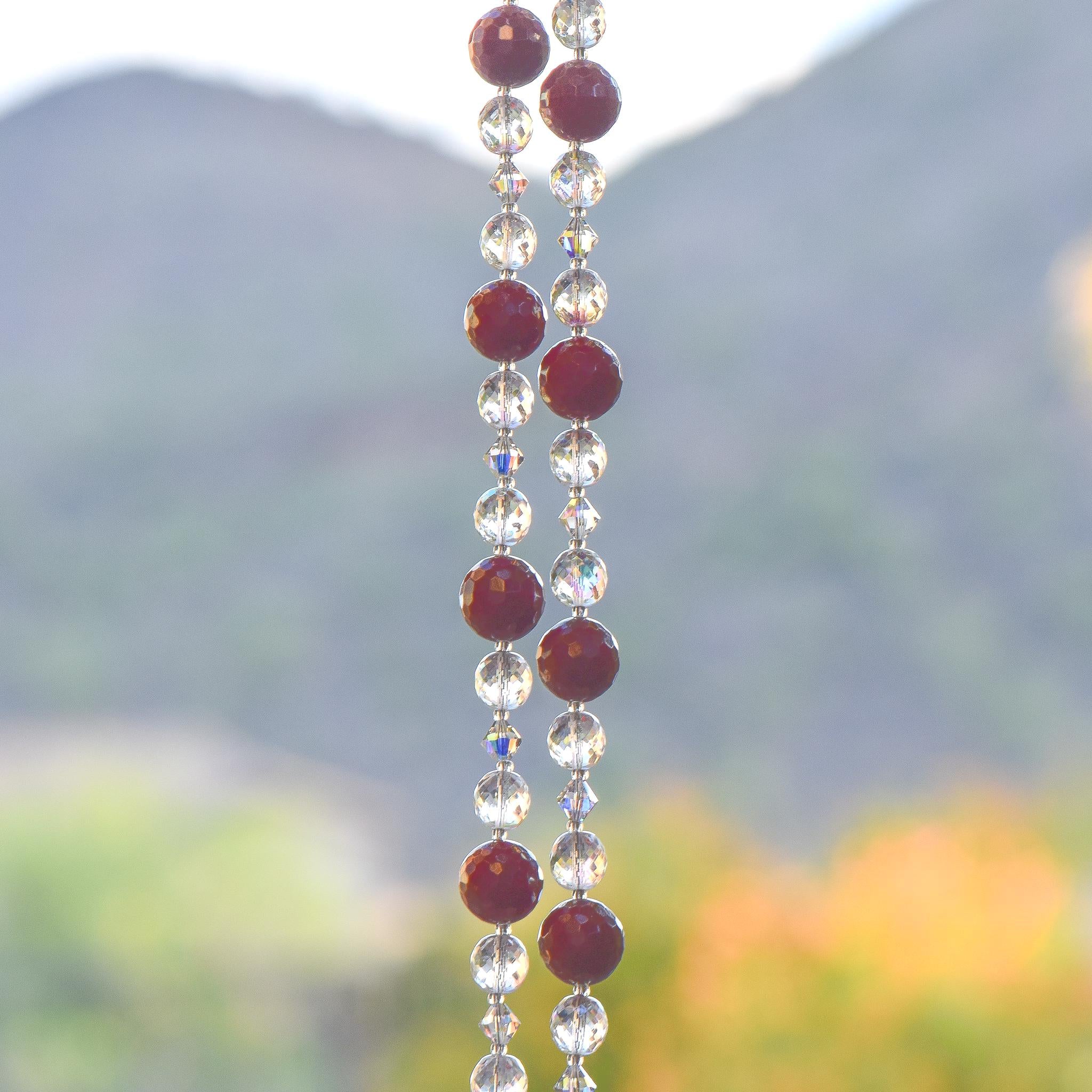 Two strands of beaded wiring hanging vertically against landscape background. Each wire has dark pinkish red jade stone beads flanked by clear glass and glass crystal.