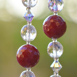 Close up of two strands of beaded wiring hanging vertically against landscape background. Each wire has dark pinkish red jade stone beads flanked by clear glass and glass crystal.