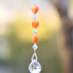 A crystal suncatcher with a clear crystal ball at the bottom and a string of orange heart-shaped beads above it.