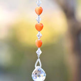 A crystal suncatcher with a clear crystal ball at the bottom and a string of orange heart-shaped beads above it.