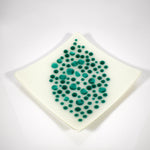 A square, off-white dish with a diamond shape. The dish is decorated with teal-colored, circular dots of varying sizes, clustered in the center. The dots create a textured, organic pattern, adding visual interest to the minimalist design. The dish has slightly rounded edges.