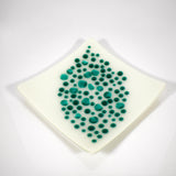 A square, off-white dish with a diamond shape. The dish is decorated with teal-colored, circular dots of varying sizes, clustered in the center. The dots create a textured, organic pattern, adding visual interest to the minimalist design. The dish has slightly rounded edges.