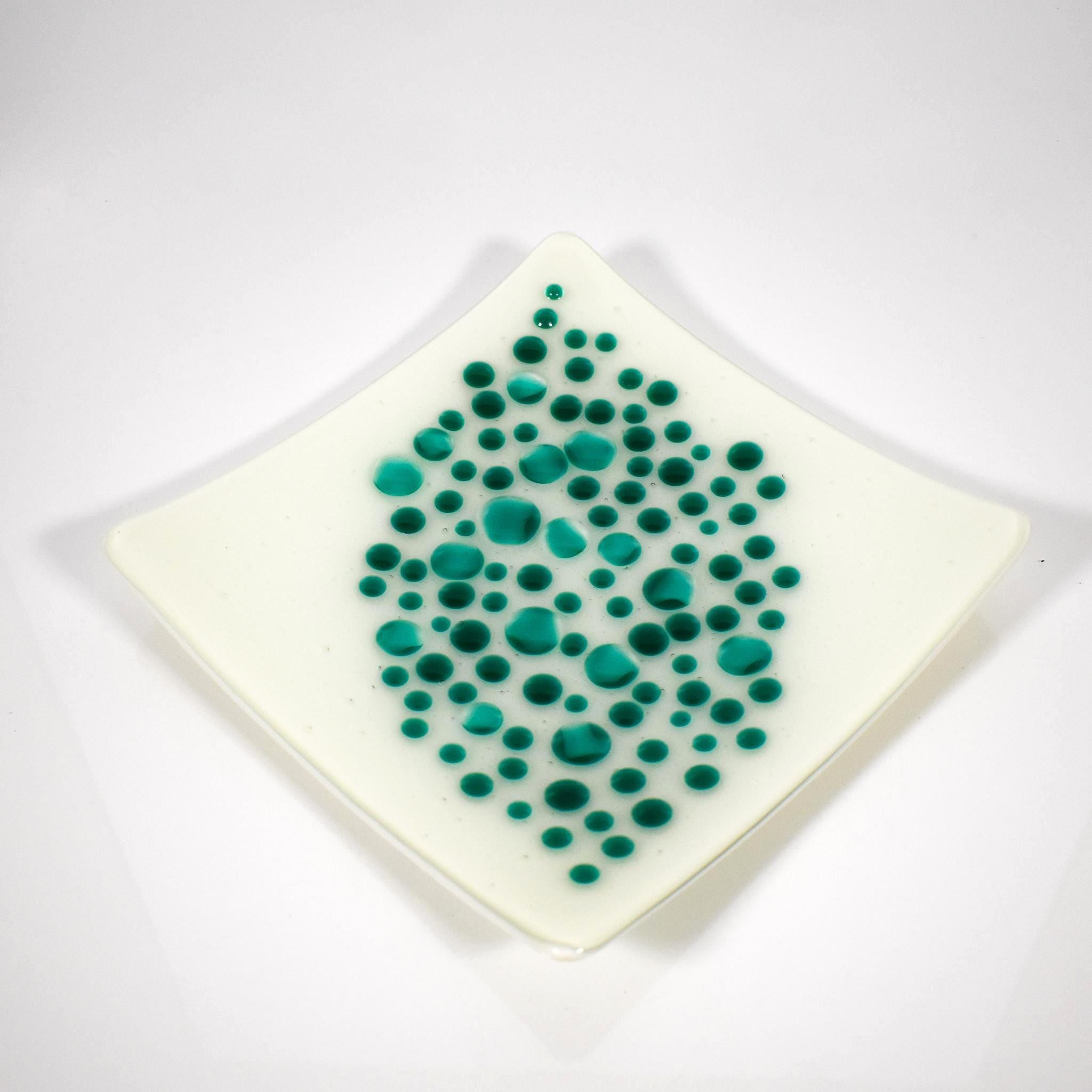 A square, off-white dish with a diamond shape. The dish is decorated with teal-colored, circular dots of varying sizes, clustered in the center. The dots create a textured, organic pattern, adding visual interest to the minimalist design. The dish has slightly rounded edges.