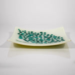Square tray made of kiln formed glass in vanilla cream with teal green, circular pieces on top in a random, diamond-shaped pattern, against white background. Viewed from the side.