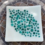 A square, white glass dish with a teal dot design. The dots vary in size and are clustered in the center of the dish. The dish is on a mosaic tile table with a circular pattern of brown, gray, and white tiles.