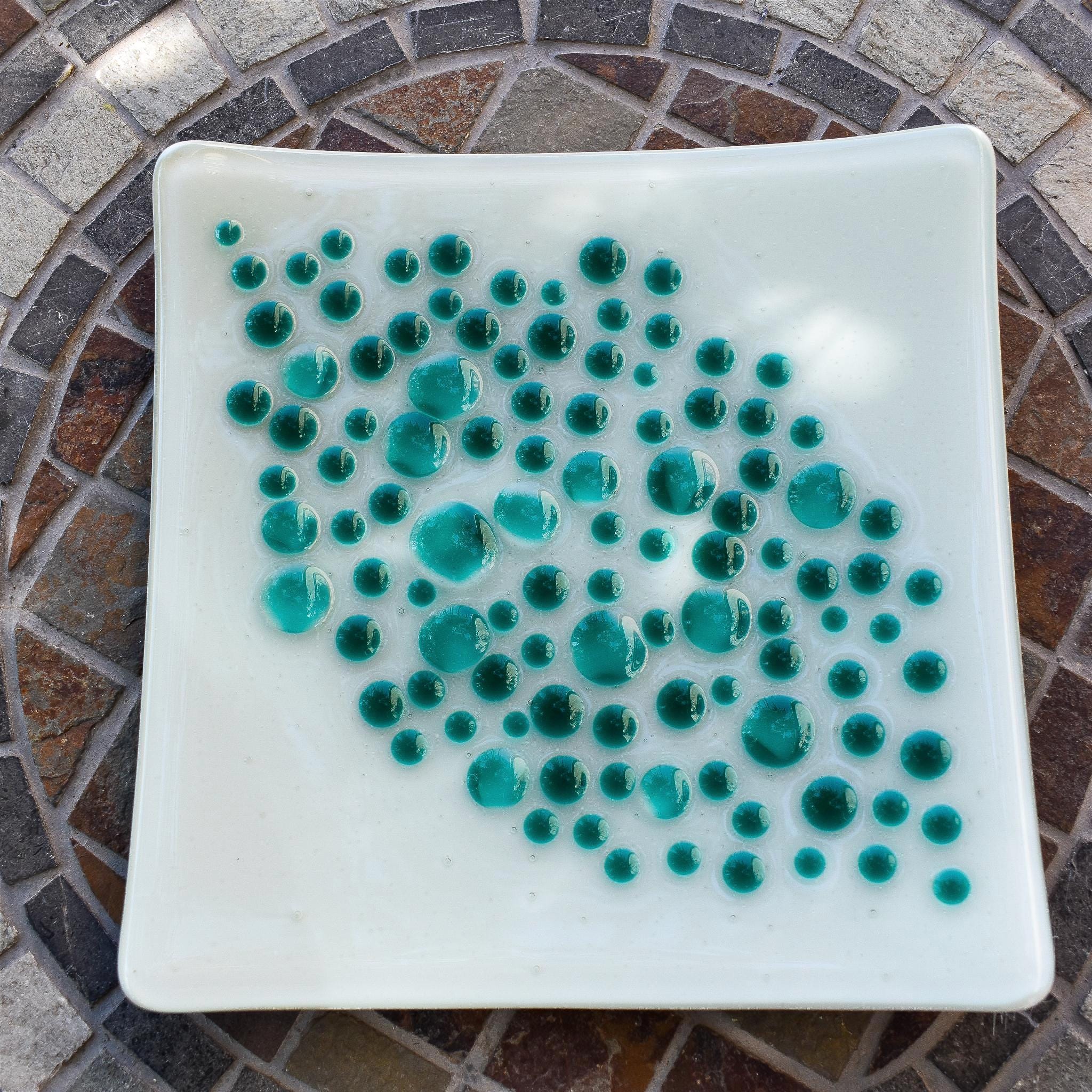 A square, white glass dish with a teal dot design. The dots vary in size and are clustered in the center of the dish. The dish is on a mosaic tile table with a circular pattern of brown, gray, and white tiles.