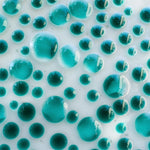 A close-up of a surface covered in teal-colored, translucent, spherical shapes of varying sizes. The spheres are densely packed, creating a bubbly, textured appearance. The background is a light, off-white color, providing contrast to the teal spheres. The image has a glossy, reflective quality.