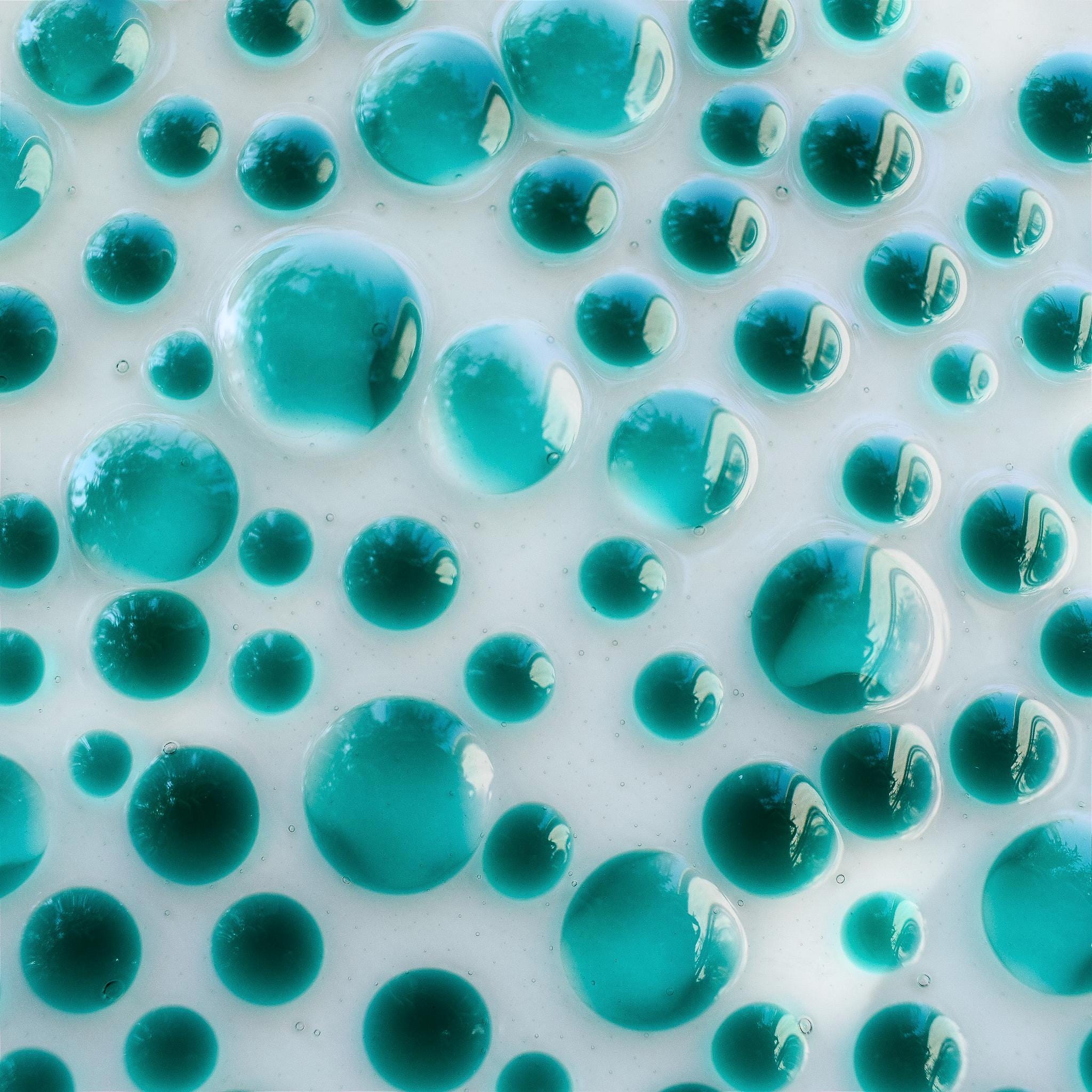 A close-up of a surface covered in teal-colored, translucent, spherical shapes of varying sizes. The spheres are densely packed, creating a bubbly, textured appearance. The background is a light, off-white color, providing contrast to the teal spheres. The image has a glossy, reflective quality.