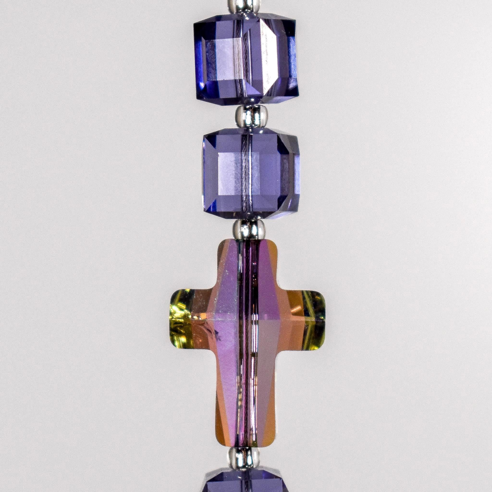 Close up of glass crystal cross beads in the color &quot;paradise shine&quot;, with two cubic purple glass beads above, against white background.