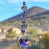 A handmade sun catcher featuring purple and iridescent crystal beads, finished with a faceted crystal butterfly pendant that reflects light in purple, blue, and gold tones. Suspended from a wire covered in silver-lined seed beads with a mountain in the background.