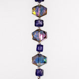 A close-up of a strand of iridescent glass beads. The beads are in a repeating pattern of a square bead followed by a hexagonal bead. The square beads are a purple color, and the hexagonal beads are a mix of purple, pink, and gold colors. White background.