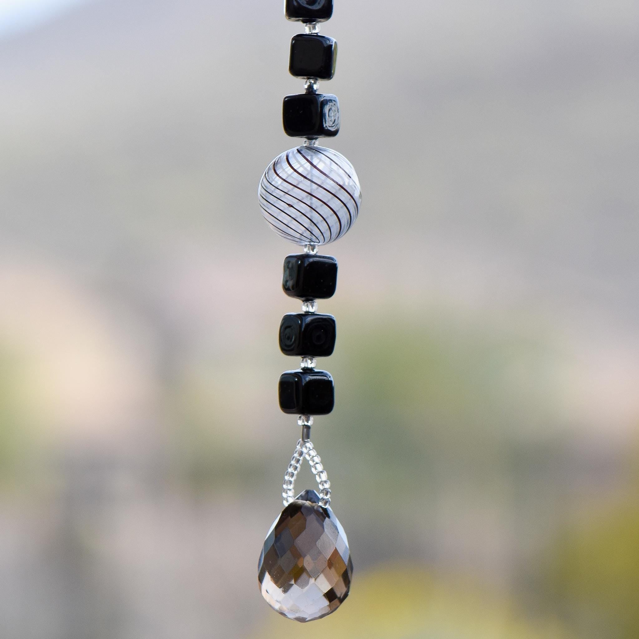 Small sun catcher hanging vertically in front of blurred background, made with six Murano glass cube beads flanking a large, round swirly bead and anchored by a large smoky quartz pendant.
