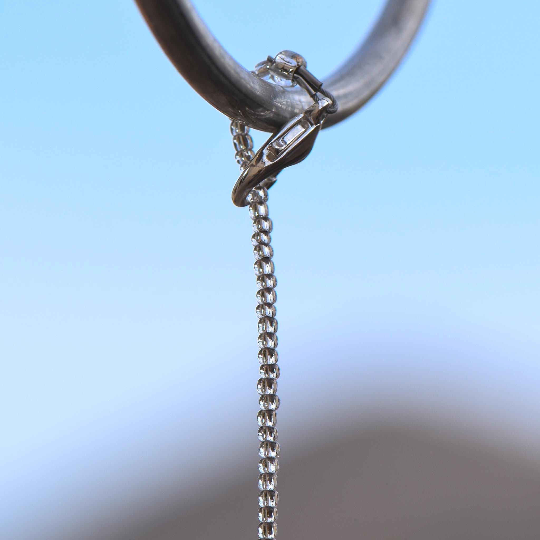 A stainless steel wire covered with silver-lined seed beads hanging from a hook against a blurred background. The top of the wire is attached to a clasp that is used to wrap the wire around the hook.