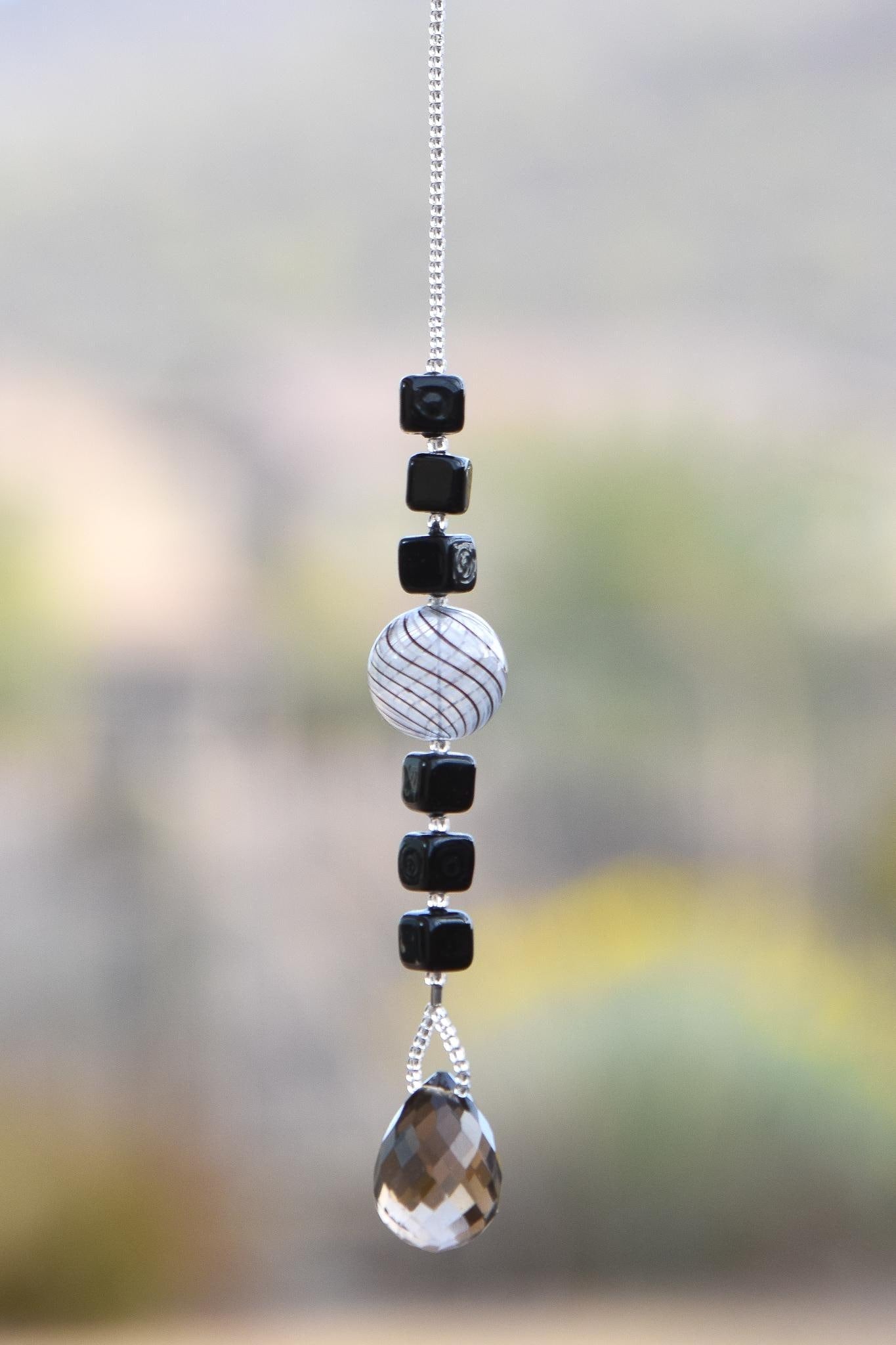 Small sun catcher hanging vertically in front of blurred background, made with six Murano glass cube beads flanking a large, round swirly bead and anchored by a large smoky quartz pendant.