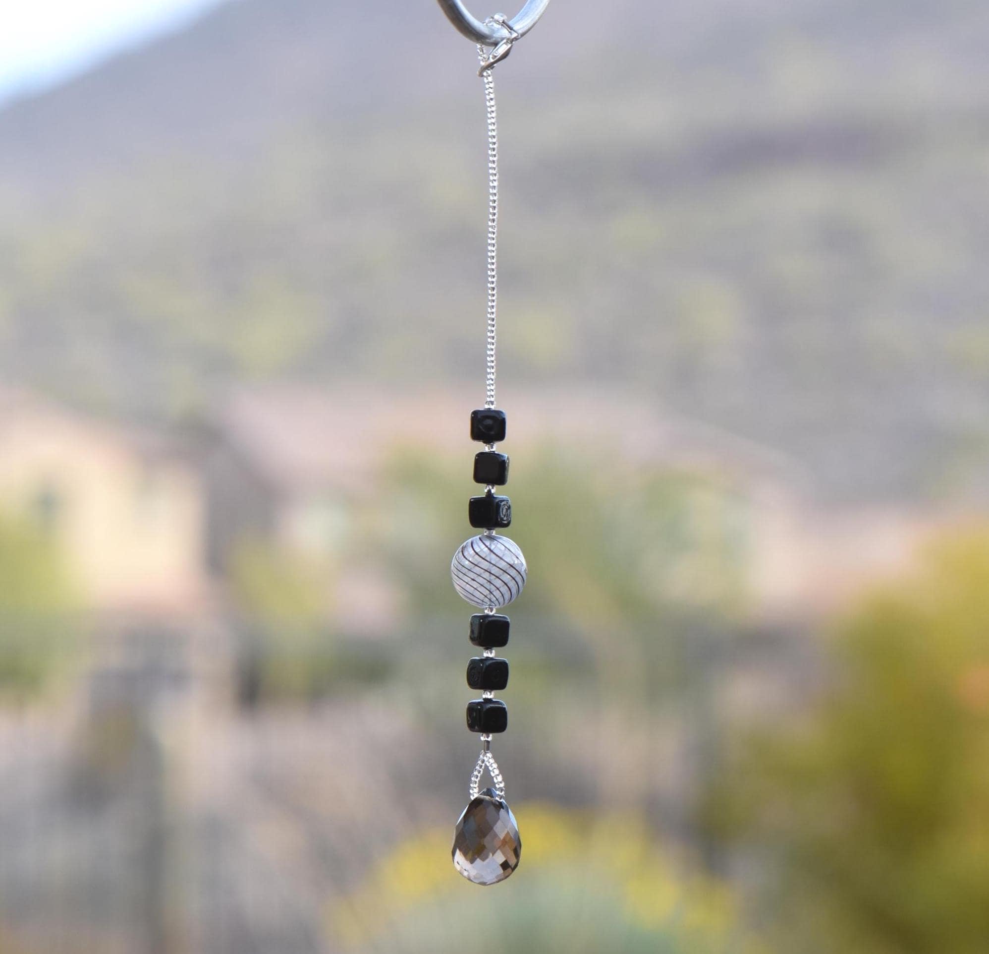 Small sun catcher hanging vertically in front of blurred background, made with six Murano glass cube beads flanking a large, round swirly bead and anchored by a large smoky quartz pendant.